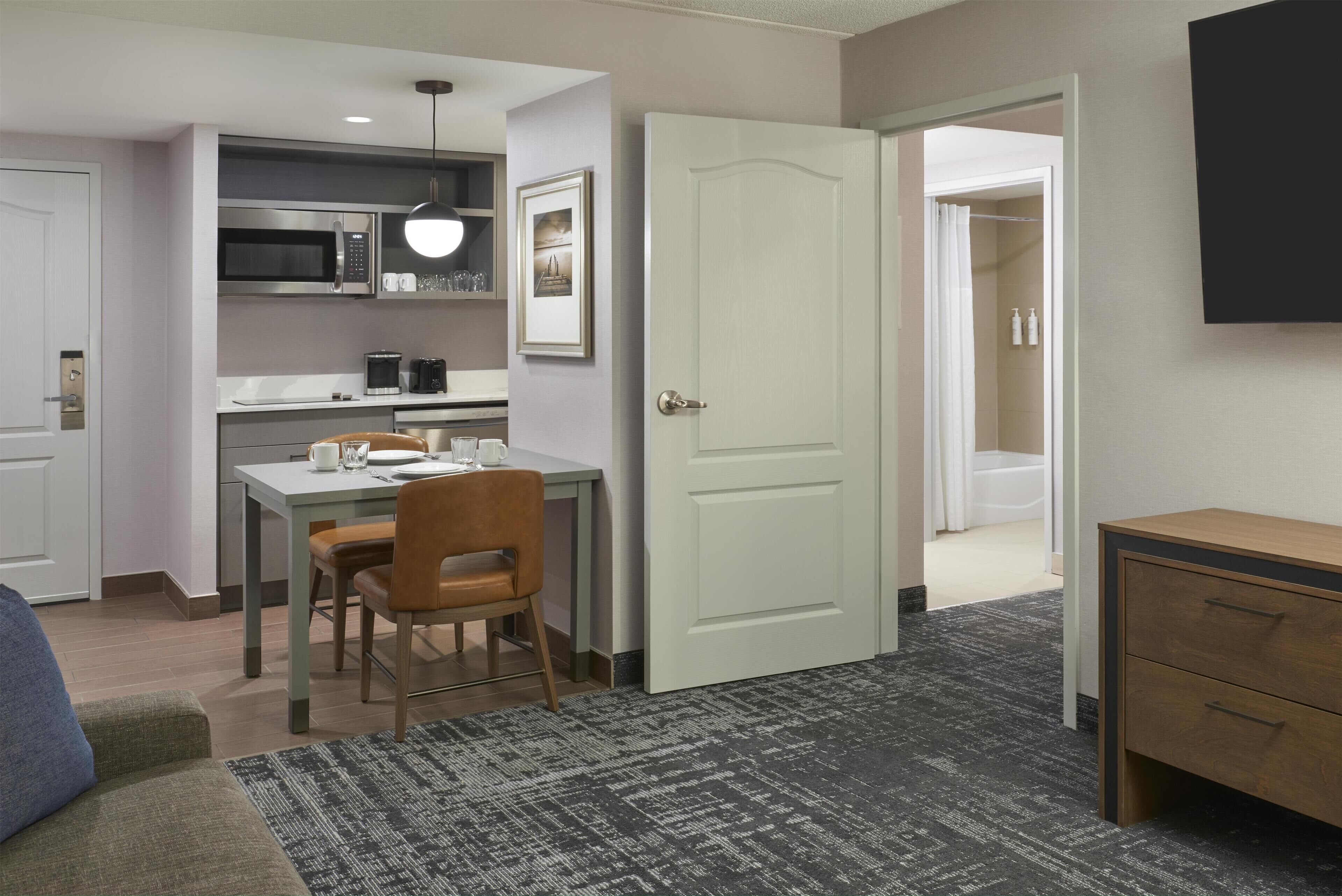 A compact hotel room with a kitchenette, a dining area, a bathroom, and a small desk.