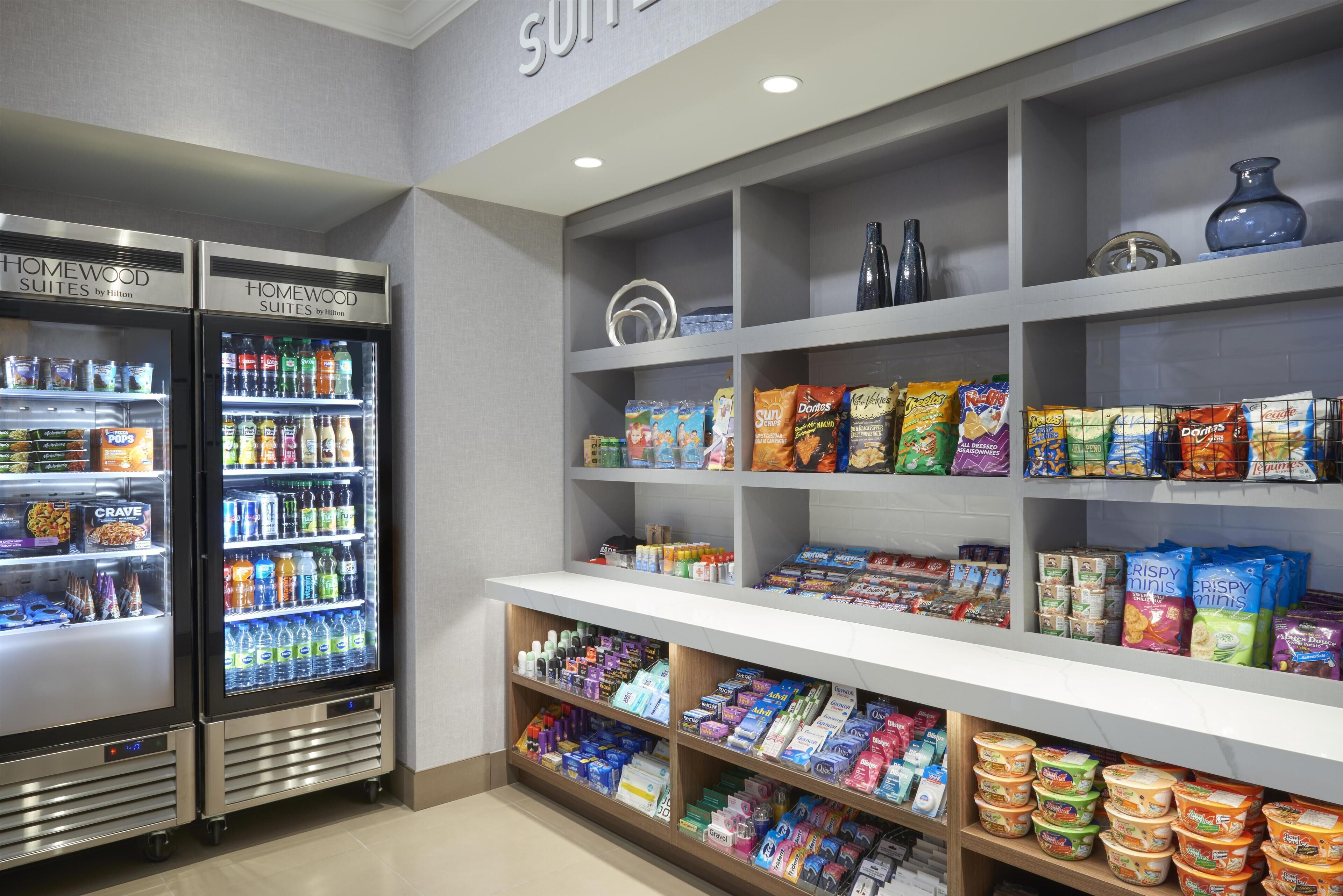 Et område til hotellets servering med to kølere til drikkevarer, hylder fyldt med snacks og et udvalg af emballerede varer.