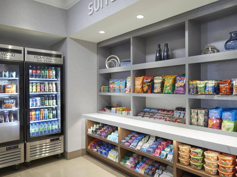 Un área de servicio de alimentos en un hotel, con dos refrigeradores para bebidas, estantes llenos de aperitivos y una variedad de productos envasados.