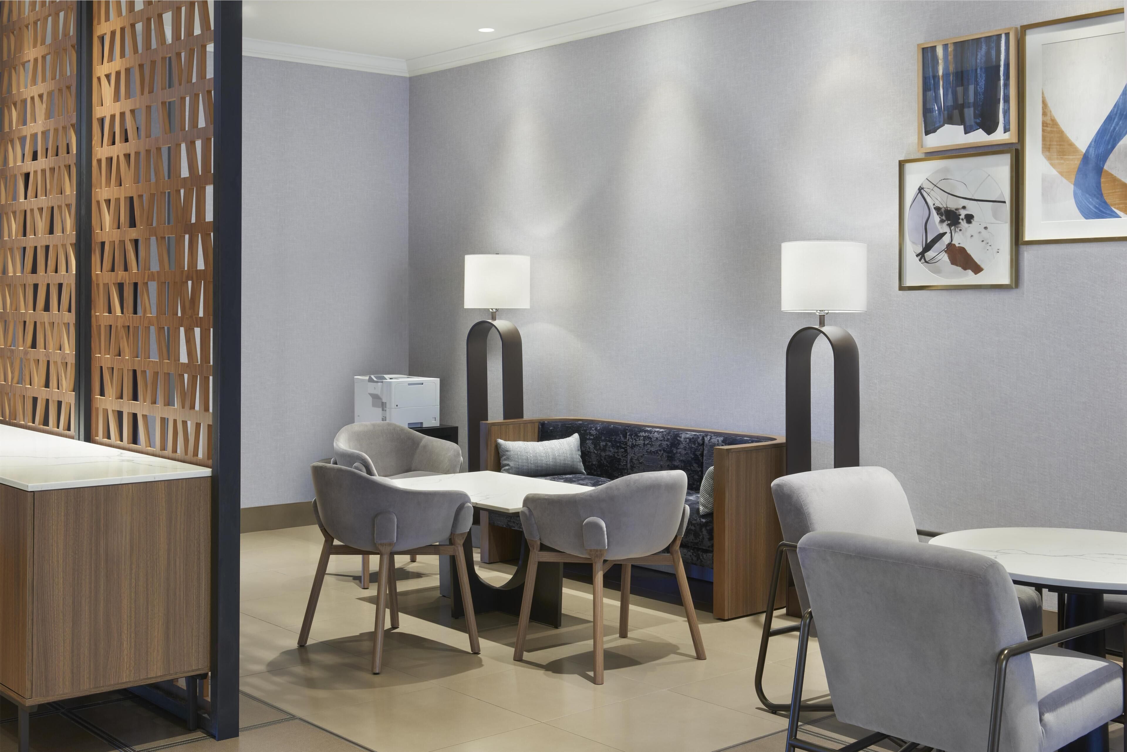 A modern hotel room with a wooden desk, two grey armchairs, a sofa, and two floor lamps.