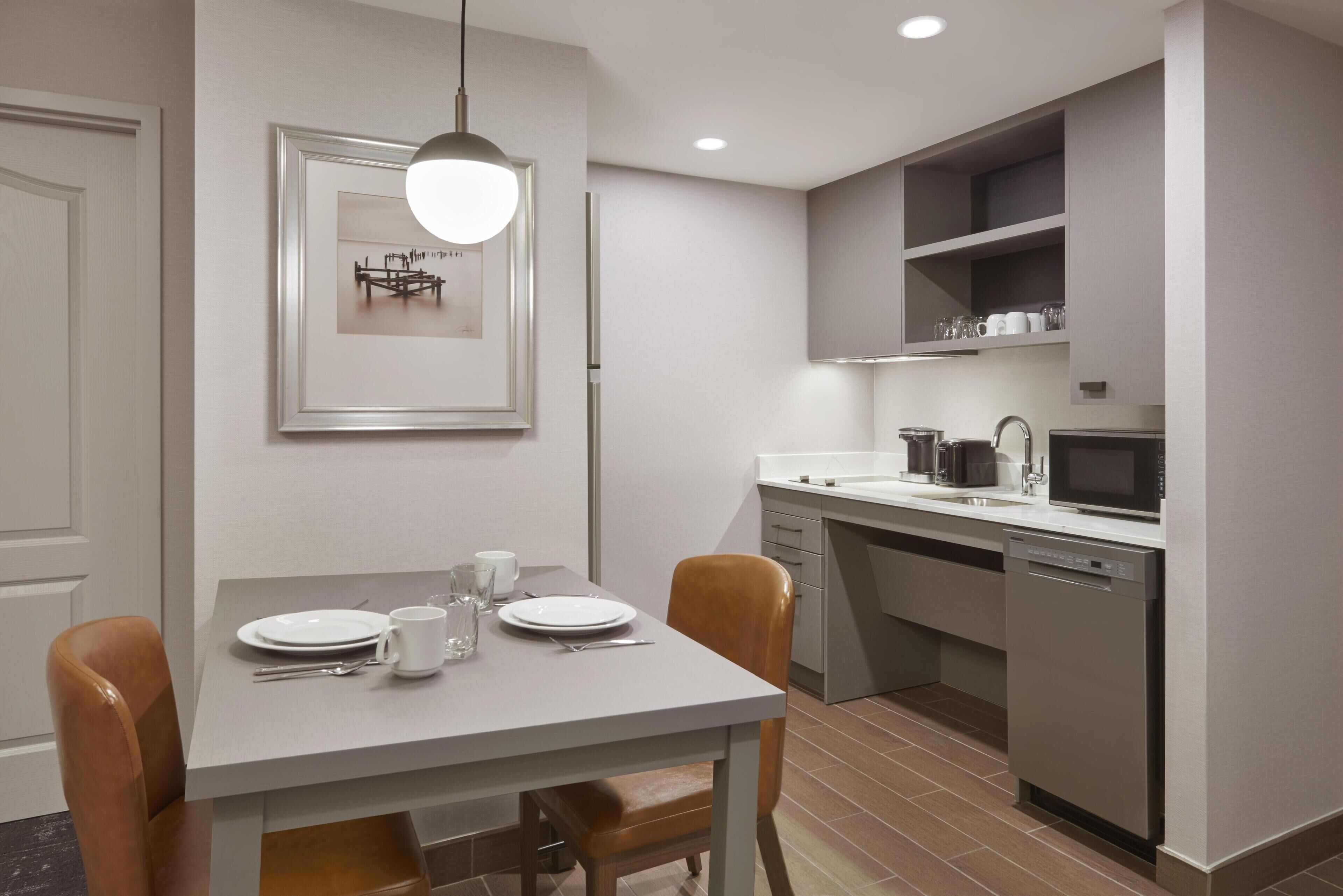 A modern kitchen with a dining area, featuring a white table set with plates, glasses, and cutlery, a hanging light fixture, and built-in appliances.