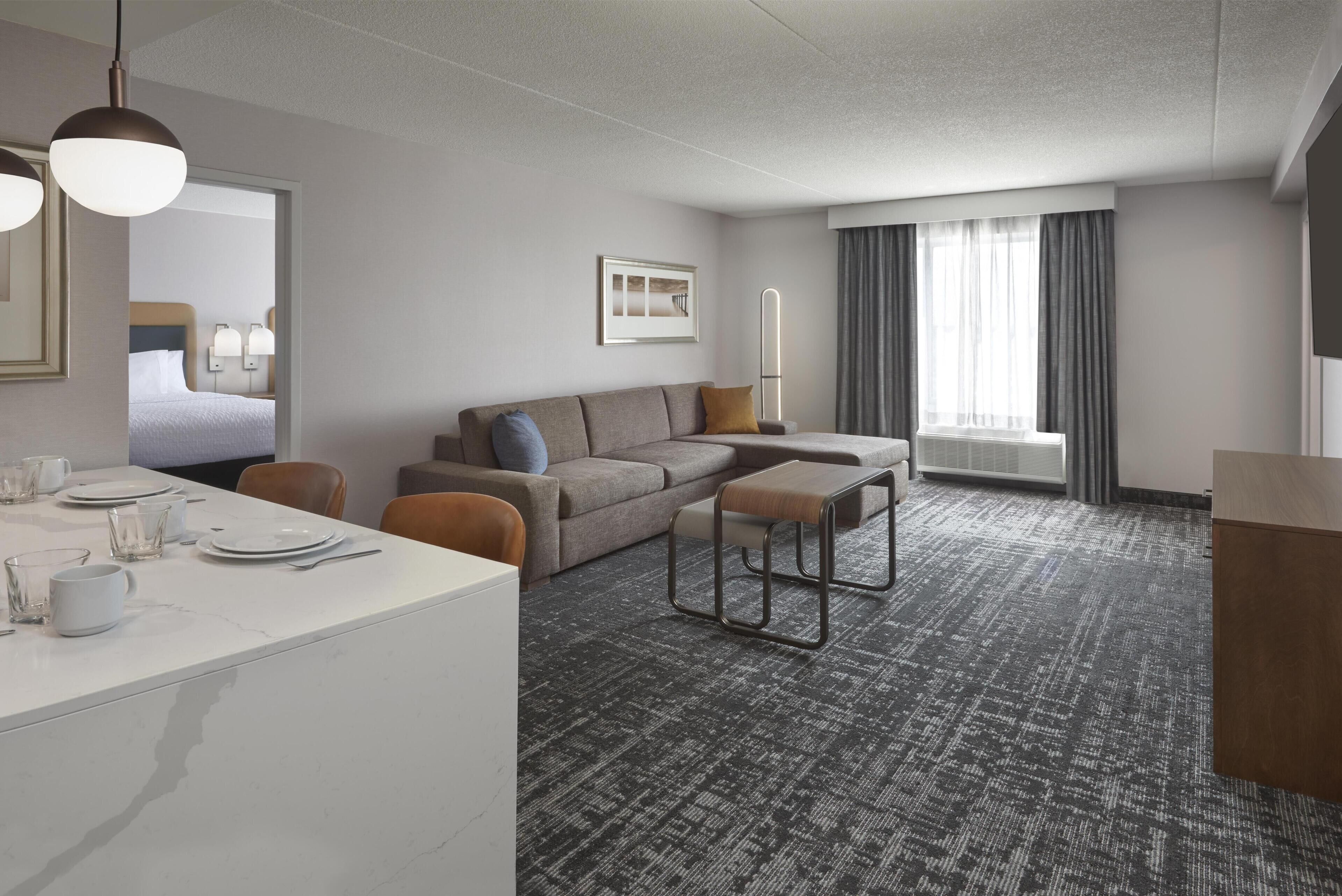 Habitación de hotel moderna con una cama grande, un sofá, una mesita y un escritorio. Cuenta con una ventana con cortinas, una lámpara de pared y un cuadro en la pared.