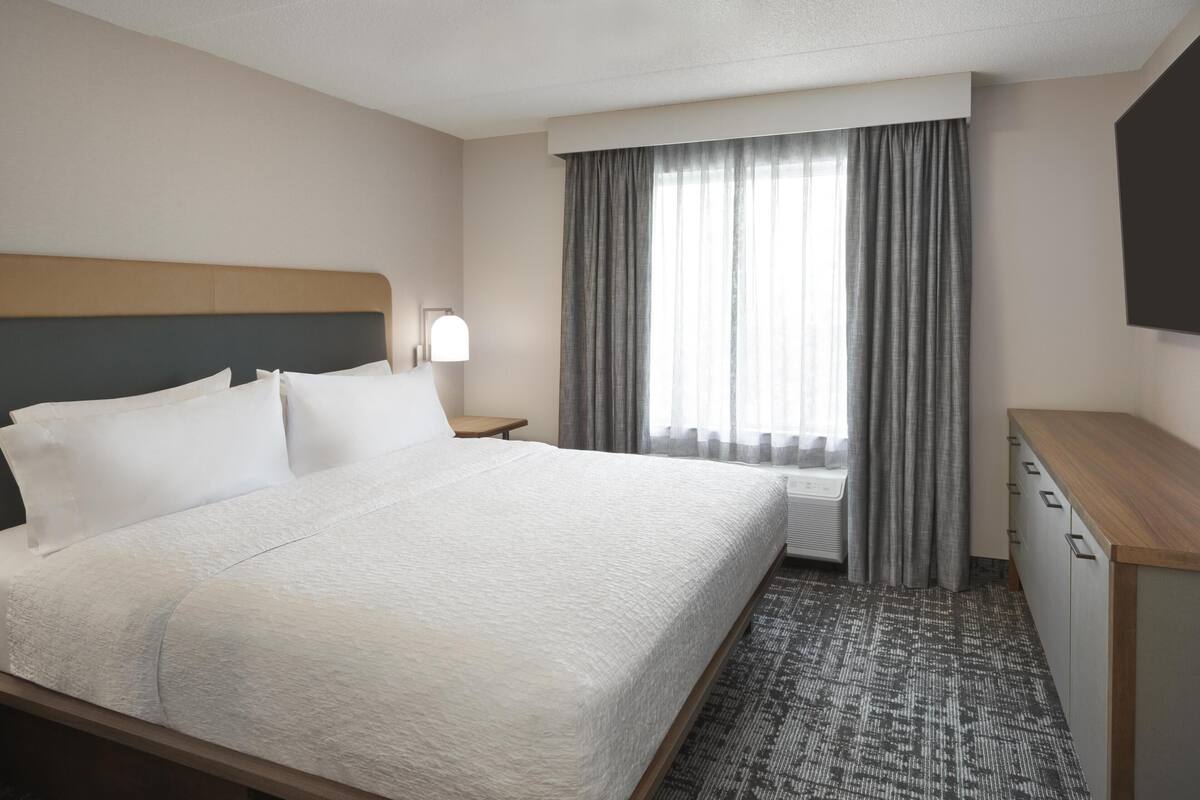 A hotel room with a large bed, a bedside table with a lamp, a wardrobe, and a window with curtains.
