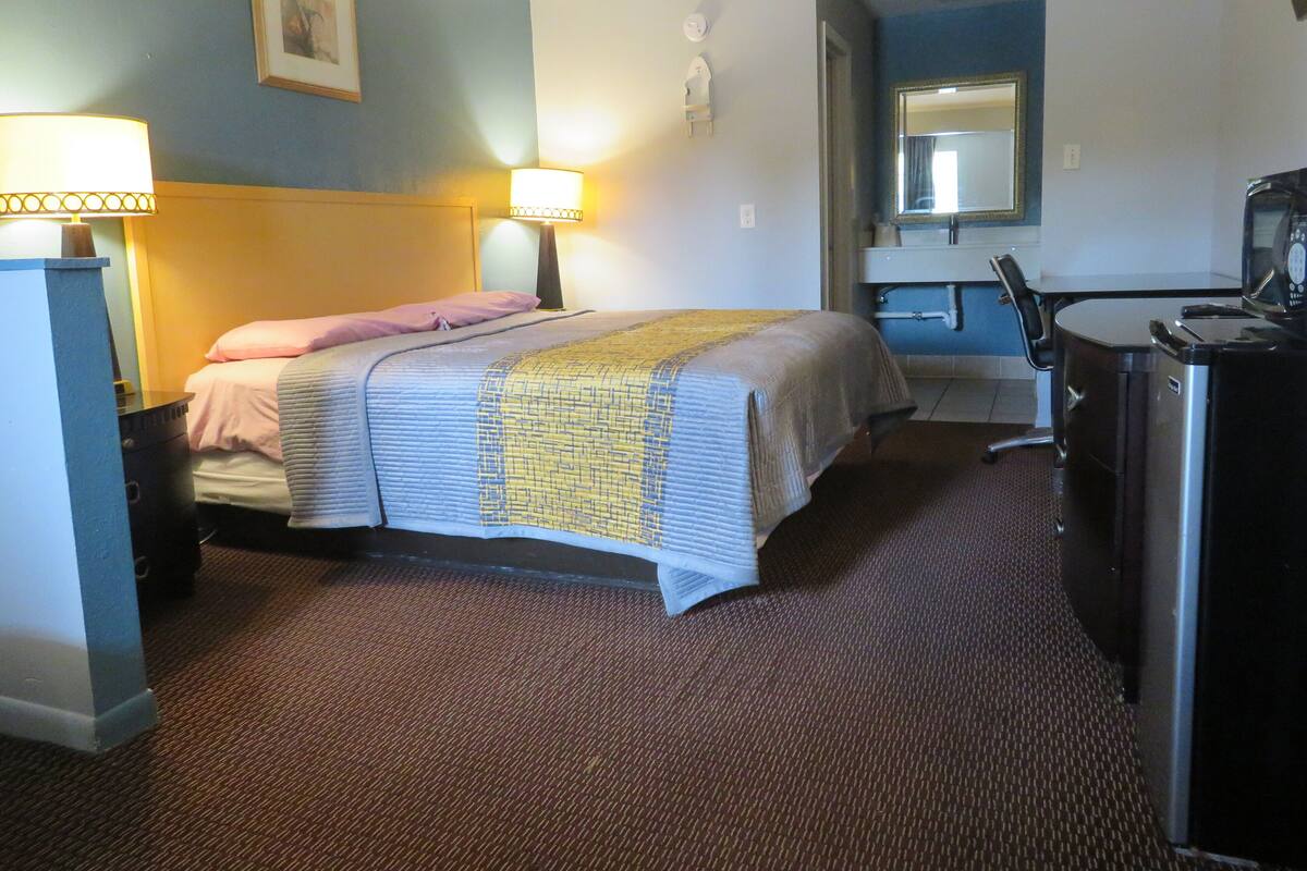 A hotel room with a bed, bedside lamps, a desk with a chair, a mirror, and a refrigerator.