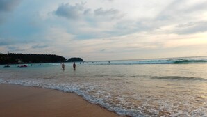 A beach with waves gently lapping onto the shore, a few people in the water, and a distant shoreline with buildings.