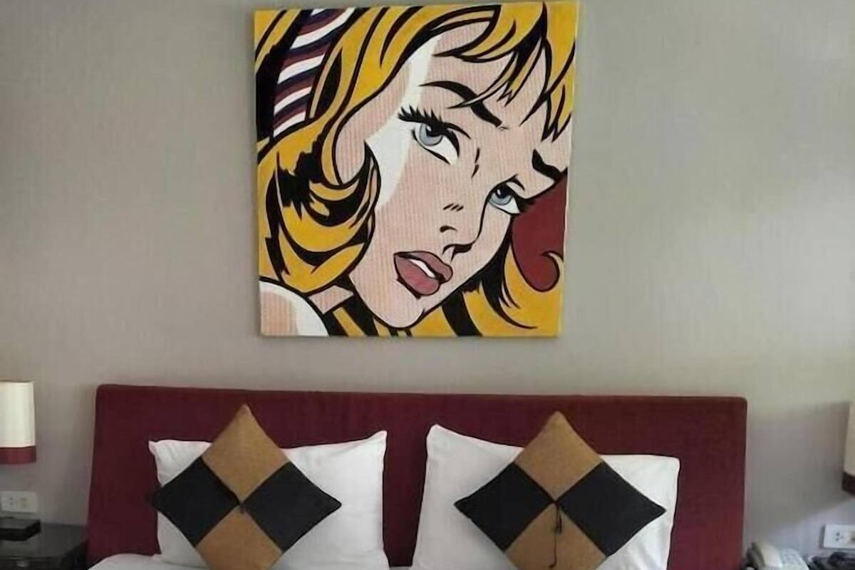 A hotel room with a bed, pillows, a painting on the wall, and a nightstand with a phone.