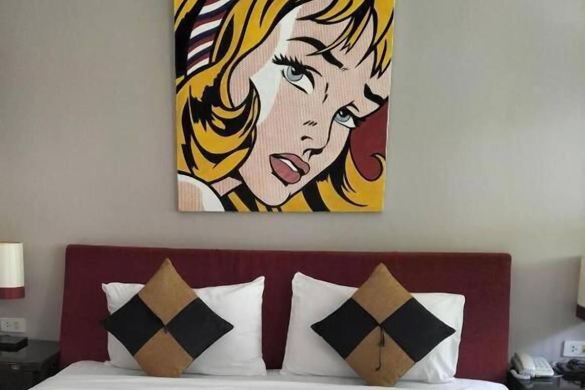 A hotel room with a bed, pillows, a painting on the wall, and a rolled towel on the bed.