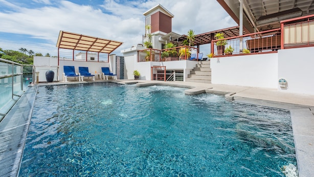 A swimming pool with blue water, surrounded by a white building with a red roof, and a covered area with blue chairs.