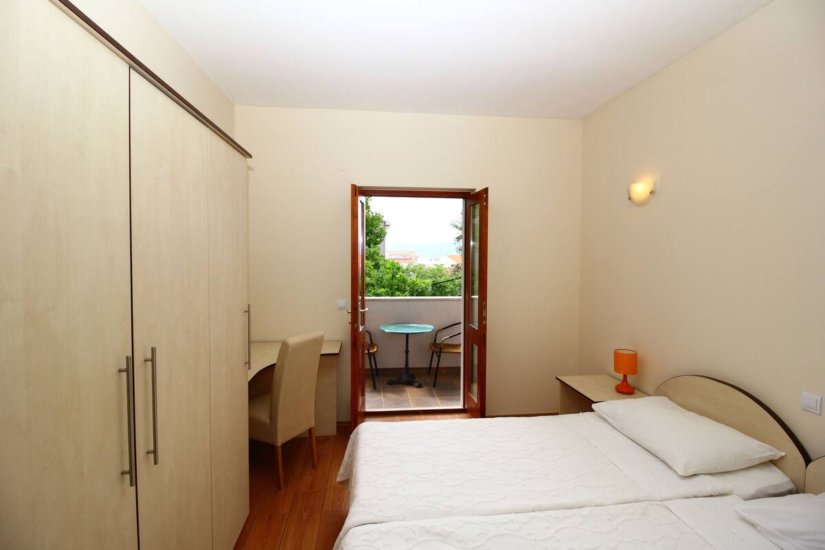 A bedroom with a bed, a desk, and a chair. There is a door leading to an outdoor area with a table and chairs.