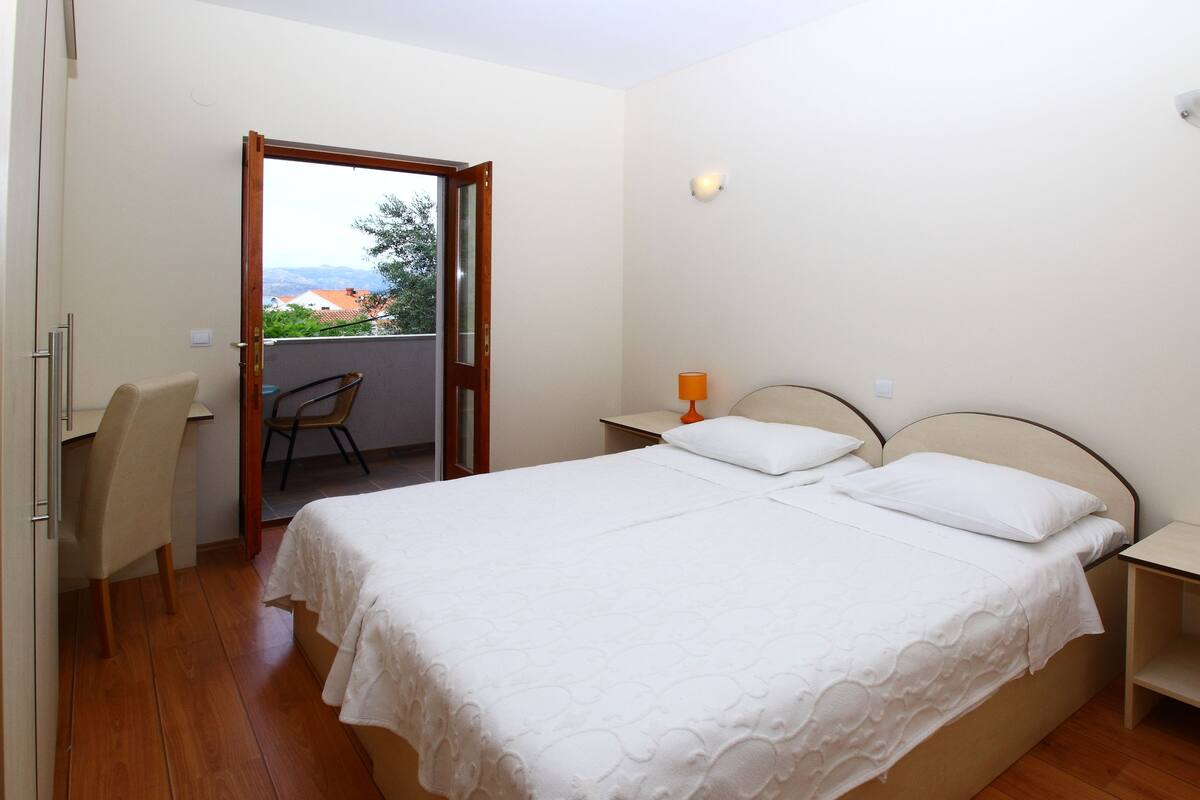 A bedroom with a double bed, a chair, a nightstand, and a view of the outdoors through an open door.