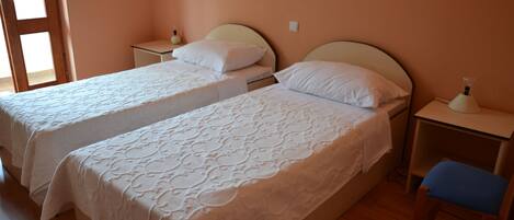 A room with two beds, wooden flooring, a nightstand with a lamp, and a chair.