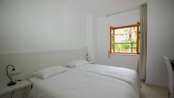 A clean, white room with a single bed, a nightstand with a lamp, and a window with a view of greenery and buildings.