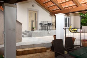 Un patio cubierto con una mesa y sillas, una escalera que lleva a una puerta, y un patinete amarillo estacionado afuera.