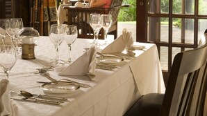 A dining table set with white tablecloth, silverware, and wine glasses, with a view of greenery outside. - Ursula's Homestead (Victoria Falls)