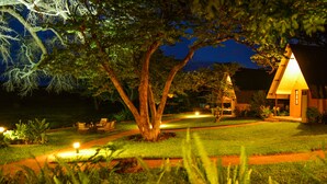 A well-lit outdoor area with a pathway leading to a building with a triangular roof. - Ursula's Homestead (Victoria Falls)