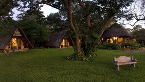 A row of thatched-roof cottages with a grassy lawn and trees in the background. - Ursula's Homestead (Victoria Falls)