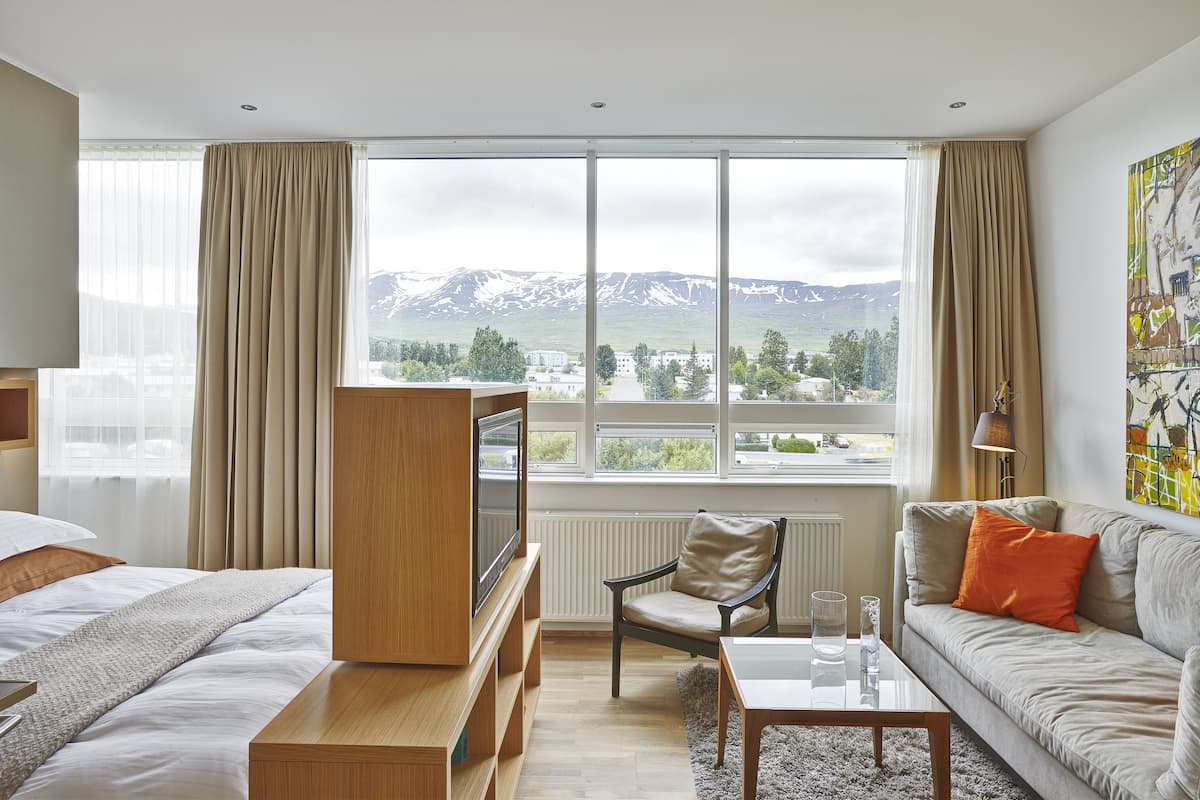 Habitación de hotel con cama, sofá, televisor y un amplio ventanal con vista a la montaña.