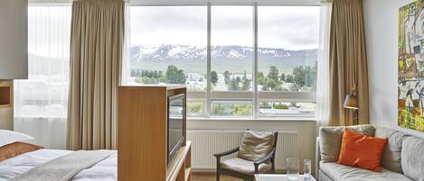 Habitación de hotel con cama, sofá, televisor y un amplio ventanal con vista a la montaña.