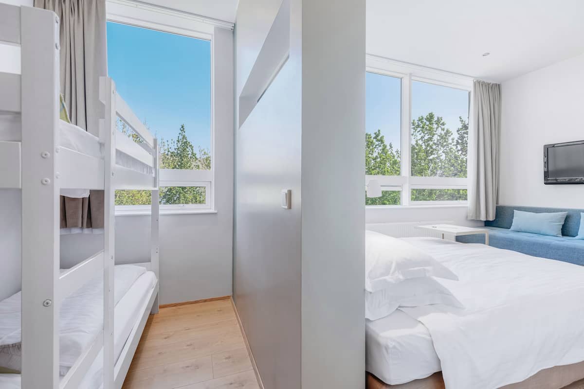 Habitación de hotel moderna con literas, cama individual, televisor y ventanas amplias con vistas a árboles.