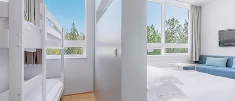 A modern hotel room with bunk beds, a single bed, a TV, and large windows offering a view of trees.