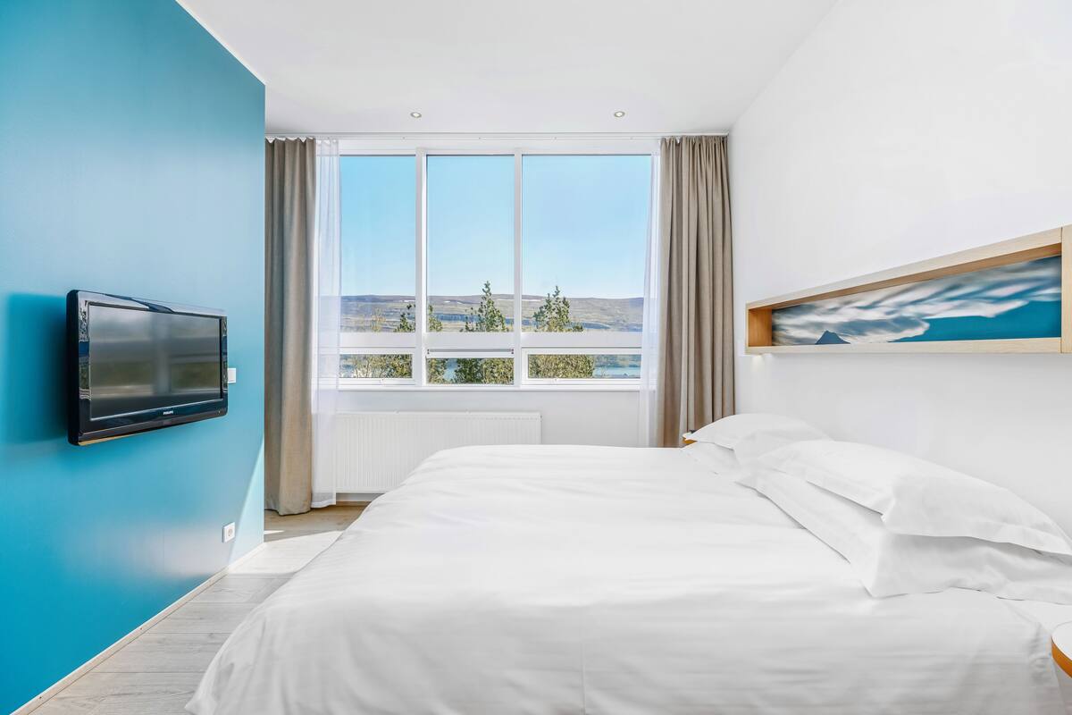 Una habitación de hotel moderna con una cama grande, un televisor de pantalla plana en la pared, un ventanal con vistas al paisaje y un cuadro colgado.