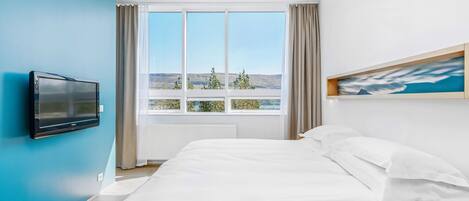 A modern hotel room with a large bed, a flat-screen TV mounted on the wall, a window with a view of the landscape, and a painting on the wall.