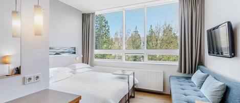 Habitació d'hotel moderna amb un llit gran, un sofà, una televisió i una finestra amb vista a arbres.
