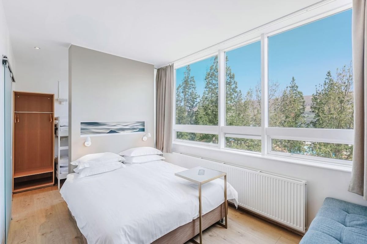 Ett sovrum med en säng, ett litet bord, en garderob och ett stort fönster med utsikt över träd.