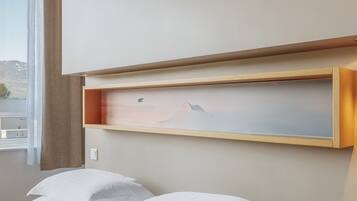 A hotel room with a bed, a wooden shelf with a painting, and a window with a view of mountains.