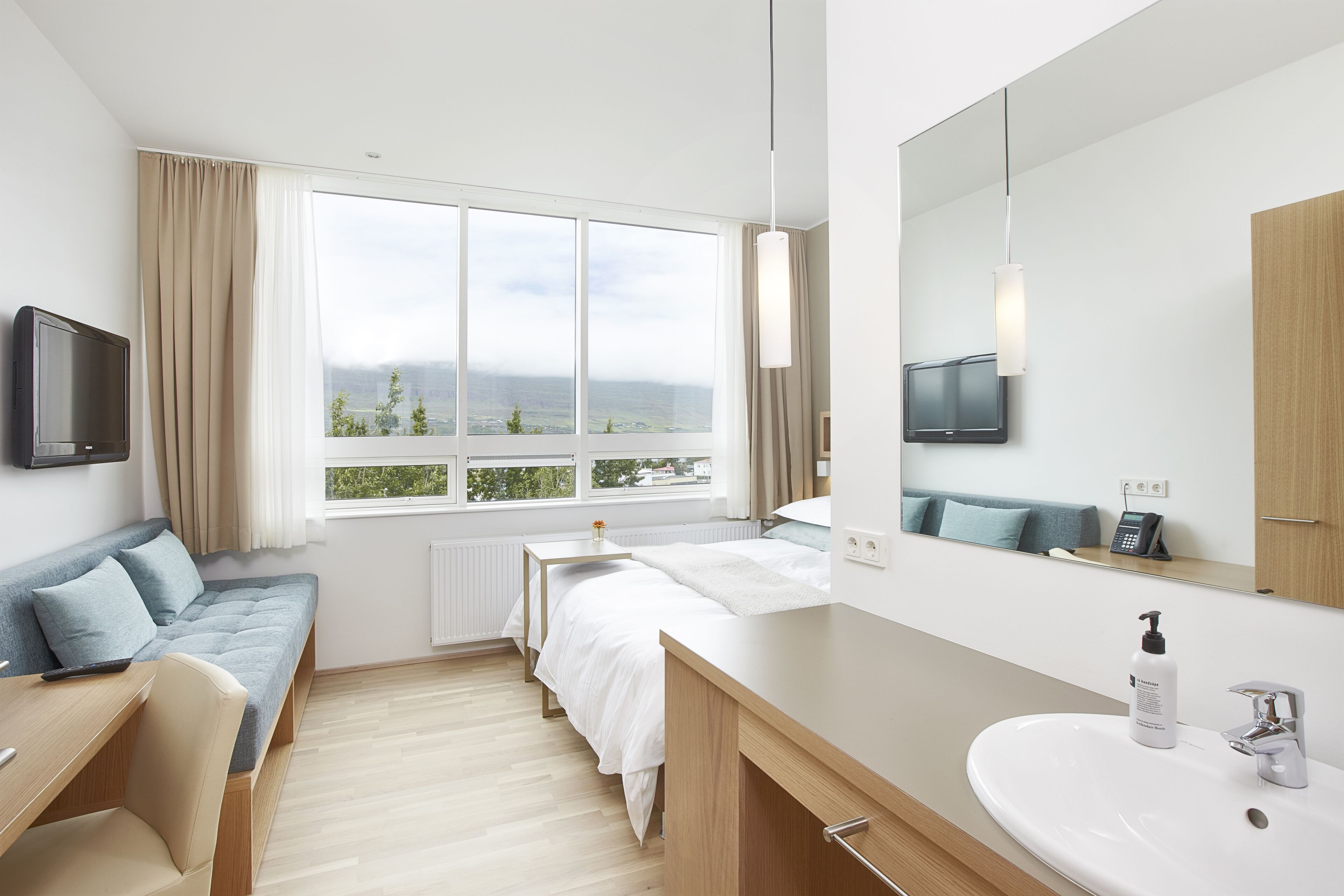 A modern hotel room with a large window, a bed, a desk, a chair, a bathroom with a sink, and a mirror.
