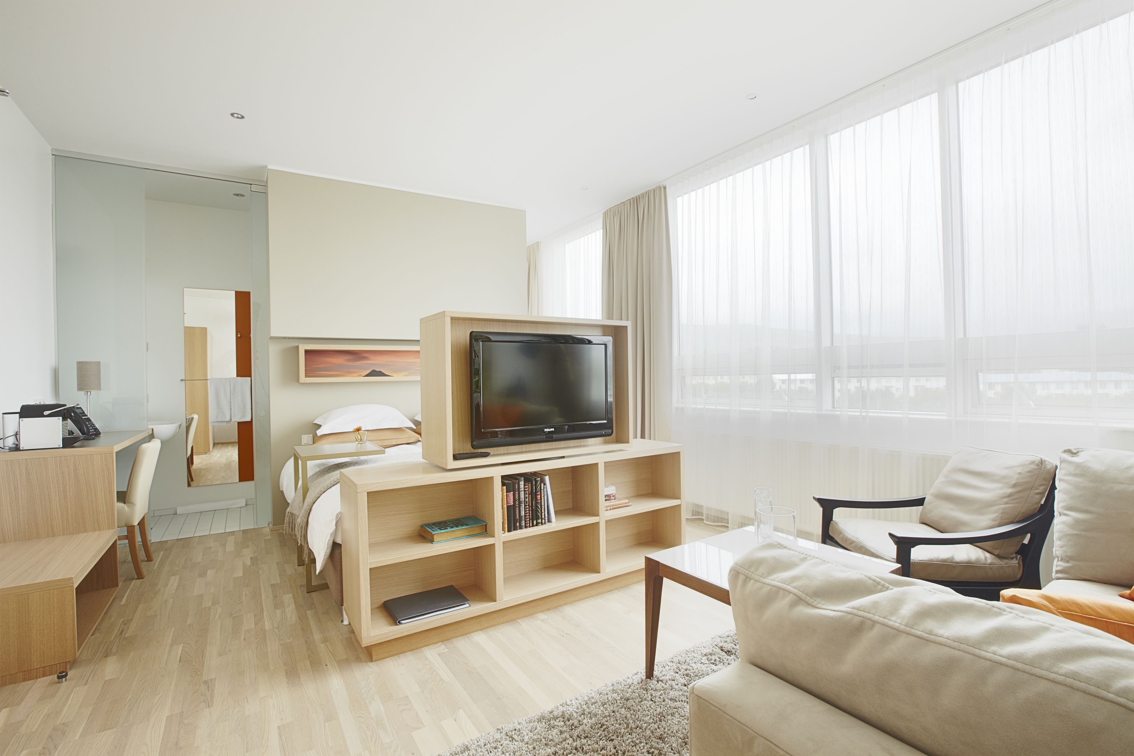 Una habitación de hotel moderna con un televisor de pantalla plana sobre un soporte de madera, un sofá, un escritorio con una silla y un ventanal con cortinas ligeras.
