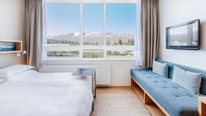 Habitación de hotel con cama, sofá azul, televisión y una ventana grande con vista a las montañas.