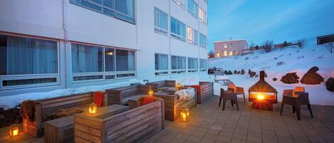 A modern outdoor seating area with wooden benches and tables, surrounded by a snow-covered landscape and a building with large windows.