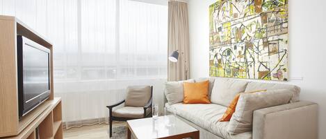 A modern living room with a flat-screen TV, a beige sofa with orange cushions, a wooden coffee table, a black chair, and a large abstract painting on the wall.