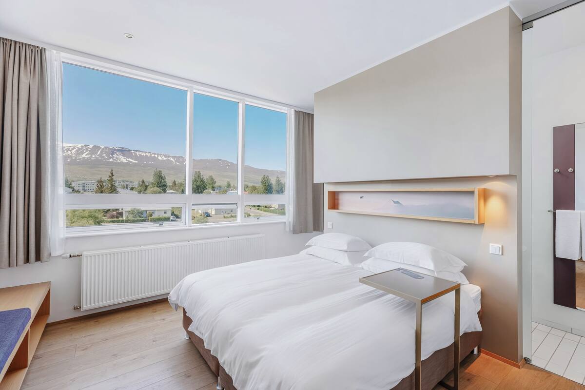 Habitación de hotel con un ventanal grande, una cama, una mesita de noche y vistas a las montañas.