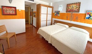Habitación de hotel con piso de madera, una cama con ropa blanca, una silla de madera y varias obras de arte en las paredes.