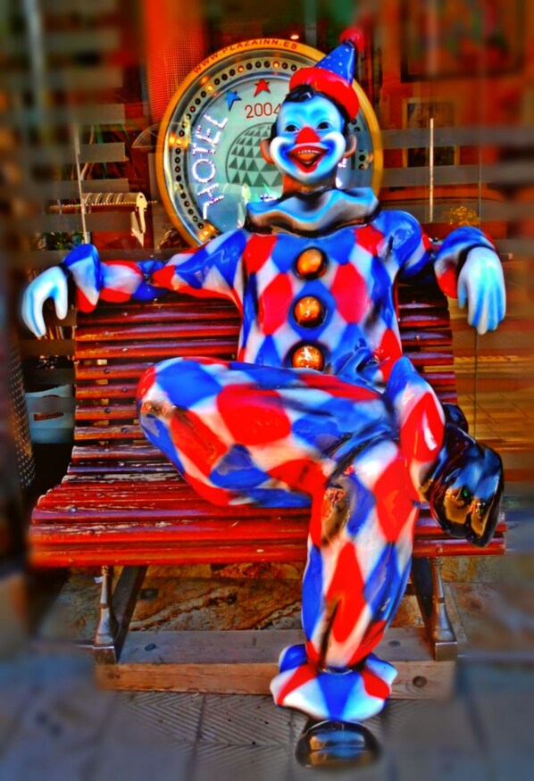 Un payaso sentado en un banco con un letrero que dice 'HOTEL' al fondo.