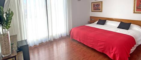 Habitación de hotel con piso de madera, cama grande con manta roja, mesita de noche, lámpara y planta en maceta.