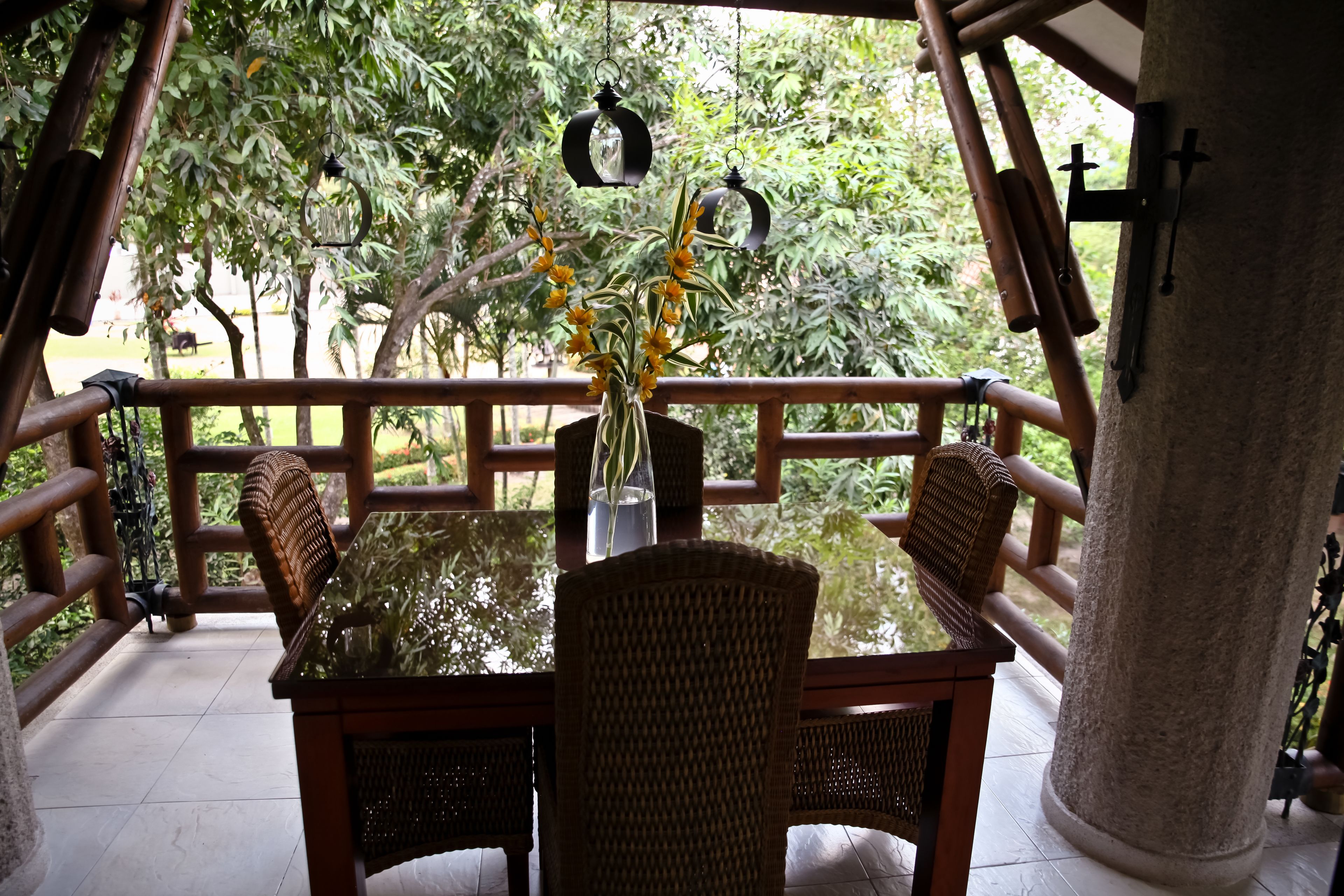 A wooden table with chairs and a vase of flowers on a balcony with a view of trees.