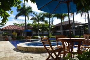 Un área de piscina con sillones de descanso y sombrillas, rodeada de palmeras y un edificio con tejado de baldosas.
