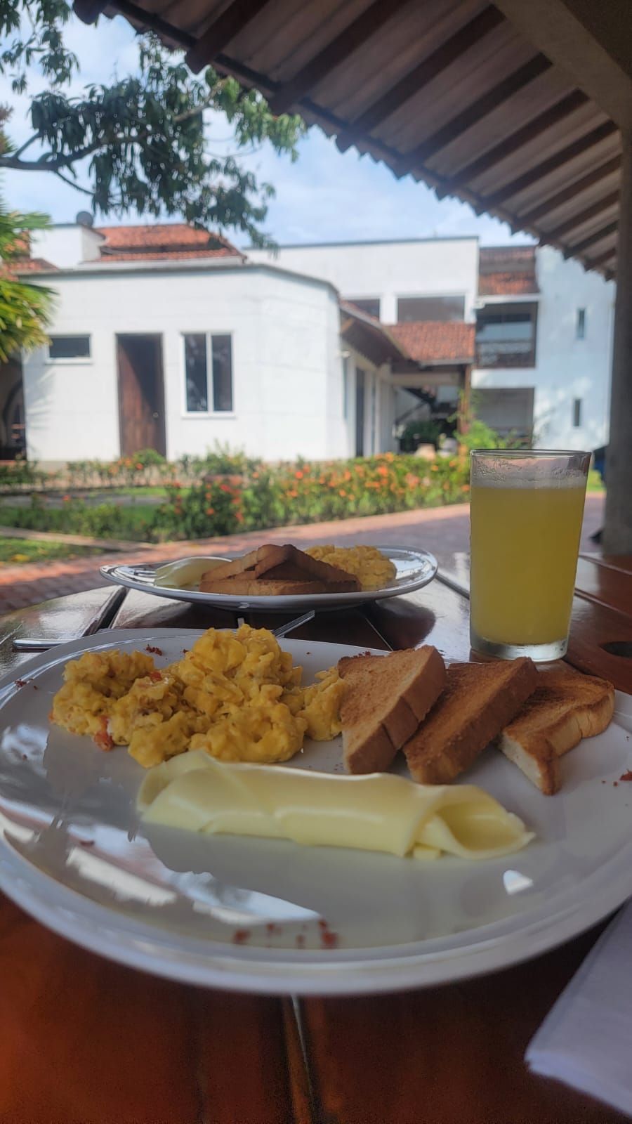 Un plat d'ous remenats, torrades i mantega damunt d'una taula, amb un got de suc, situat a l'aire lliure amb un edifici i un jardí al fons.