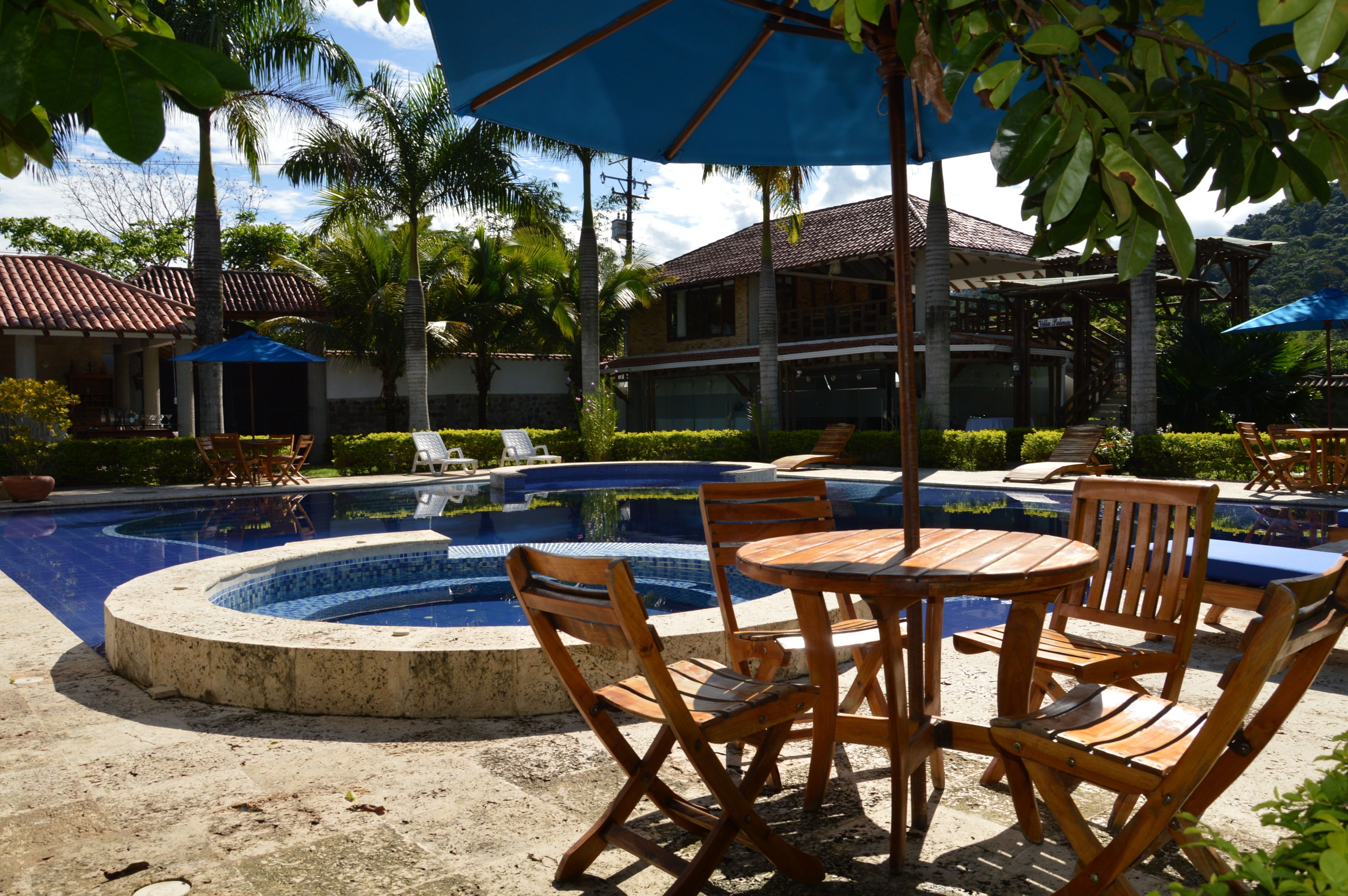 A pool area with a circular hot tub, wooden chairs, and tables under blue umbrellas.