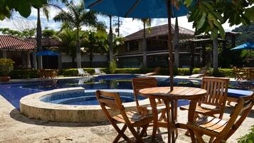 A pool area with a circular hot tub, wooden chairs, and tables under blue umbrellas.