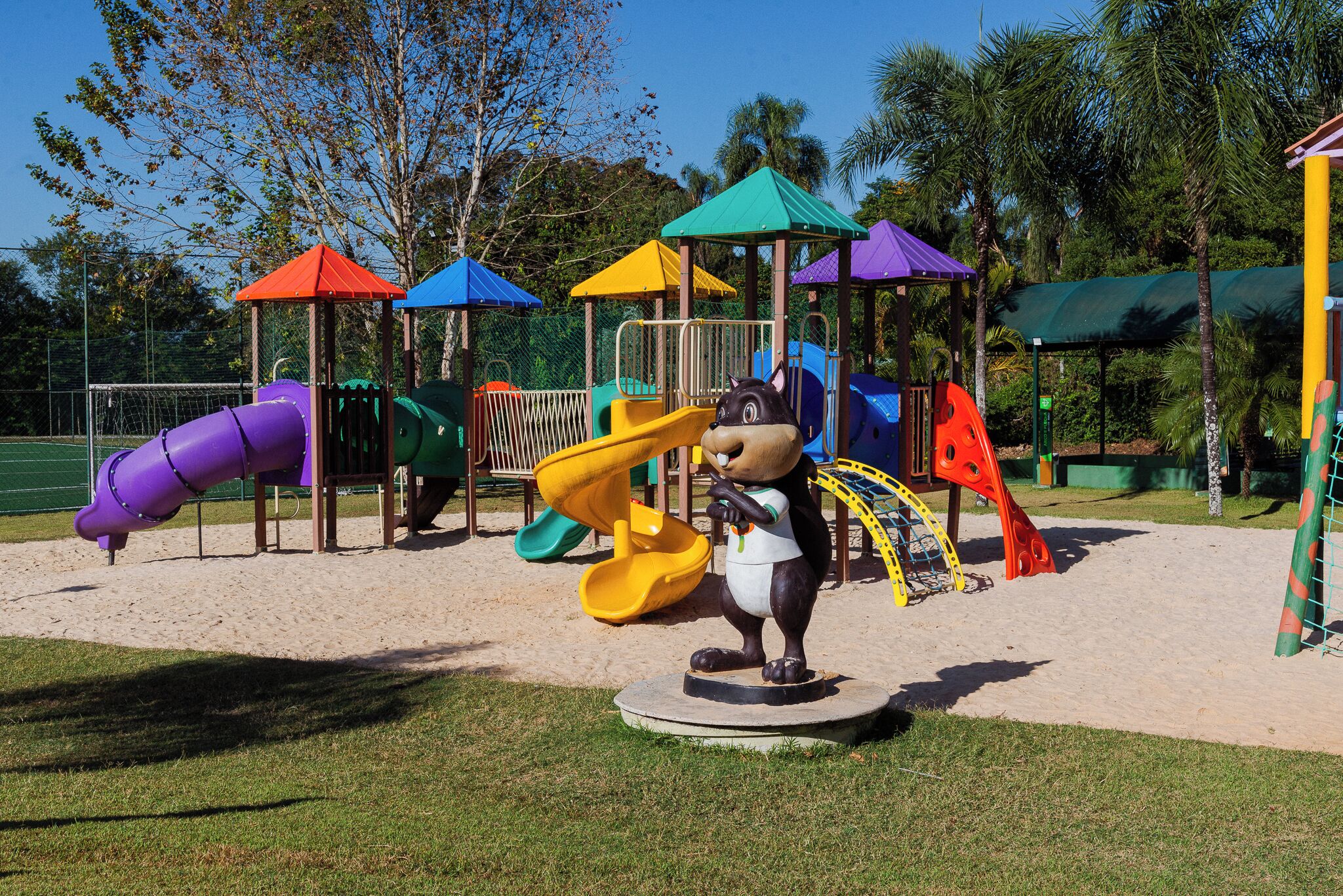 A playground with various play structures, including slides and climbing areas, surrounded by trees and a fenced area.