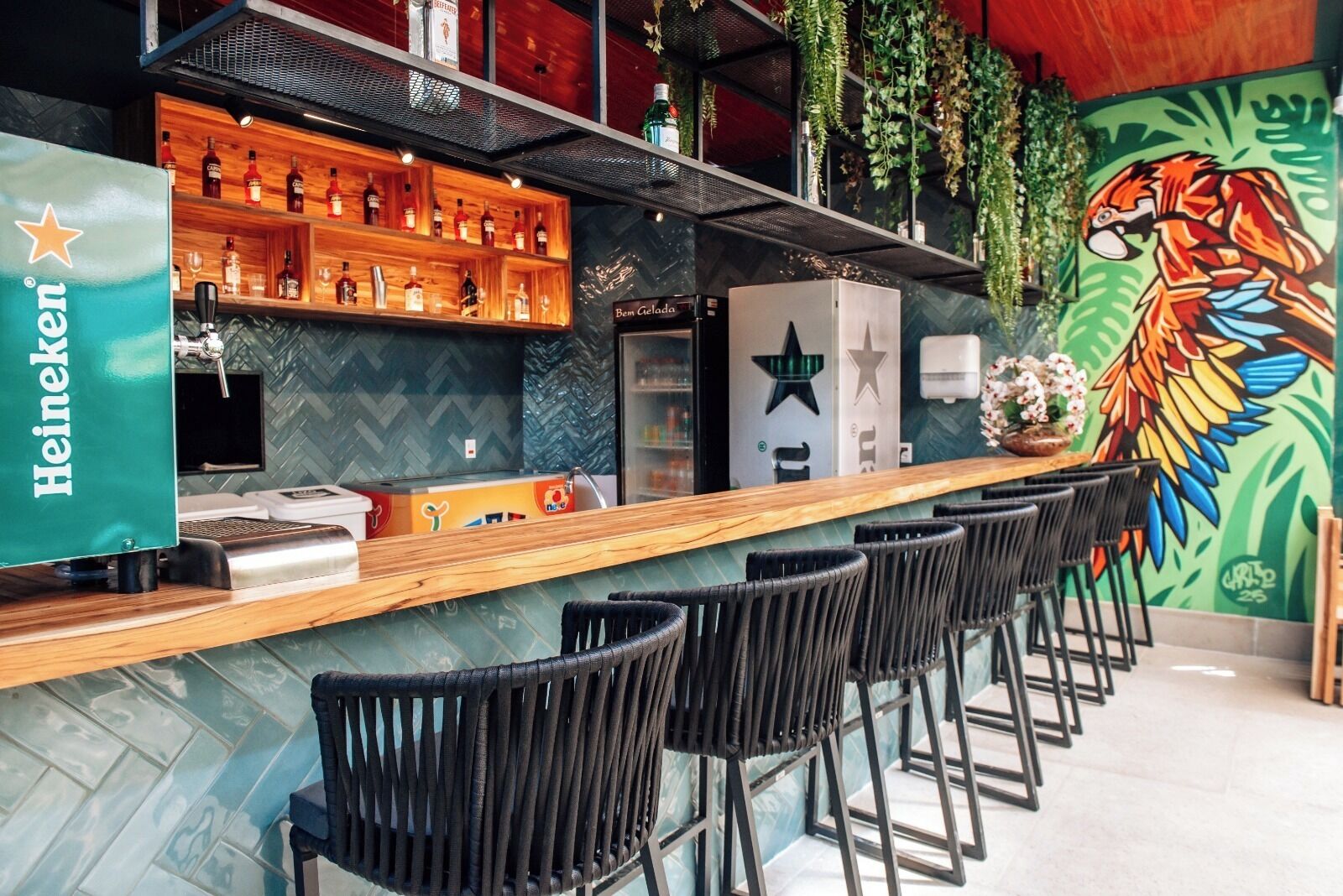 A bar area with a Heineken sign, shelves with liquor bottles, a refrigerator, and a mural of a colorful bird.