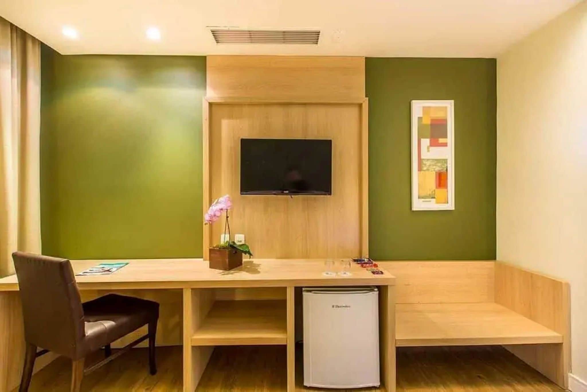 A room with a wooden desk, a television mounted on the wall, a small refrigerator, and a chair.