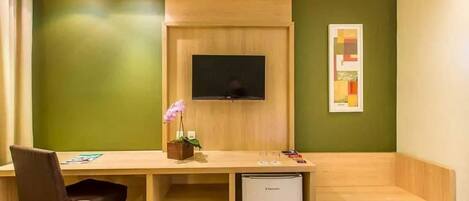 A room with a wooden desk, a television mounted on the wall, a small refrigerator, and a chair.