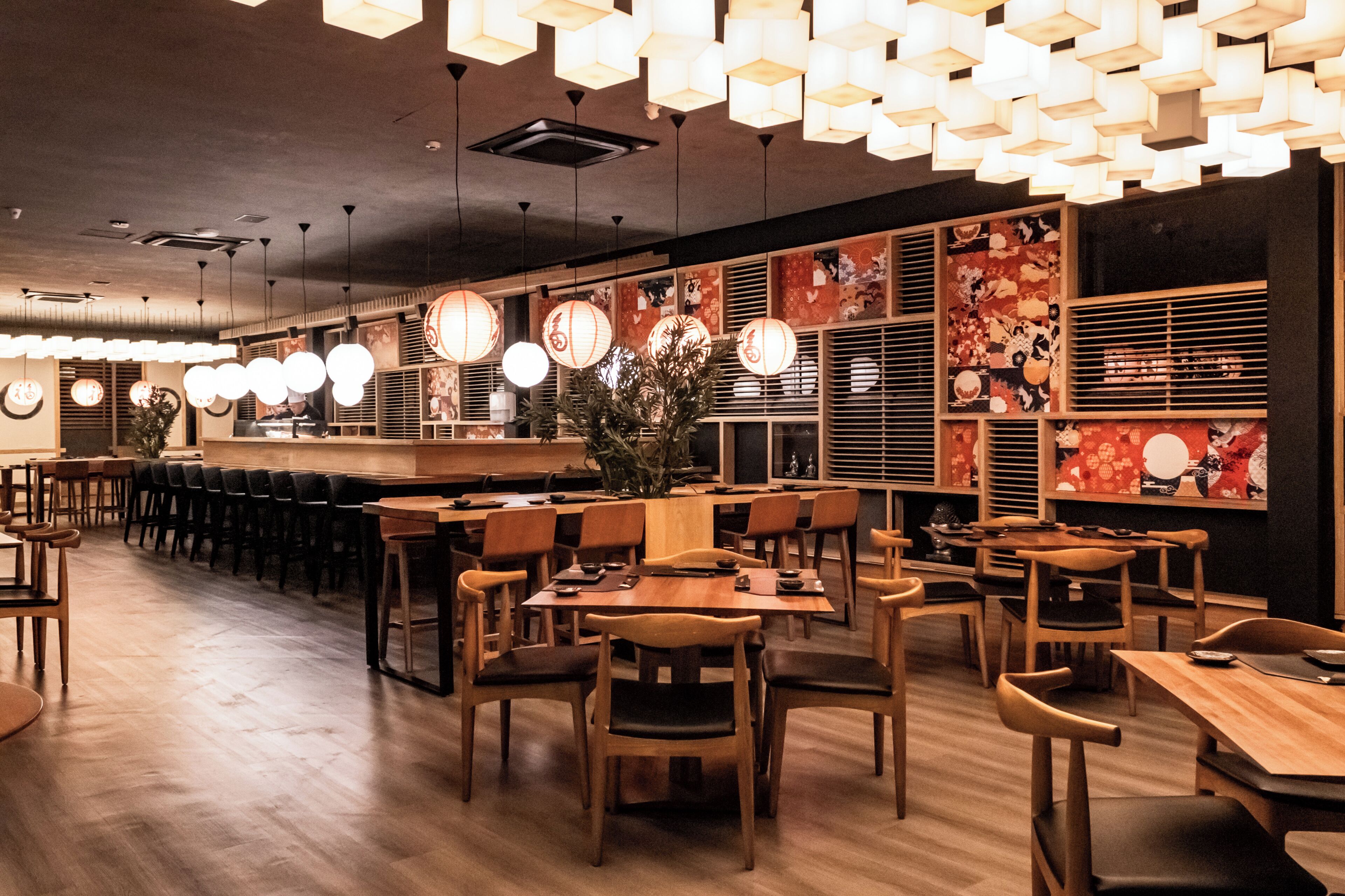 A restaurant interior with wooden tables and chairs, hanging pendant lights, and decorative wall panels.