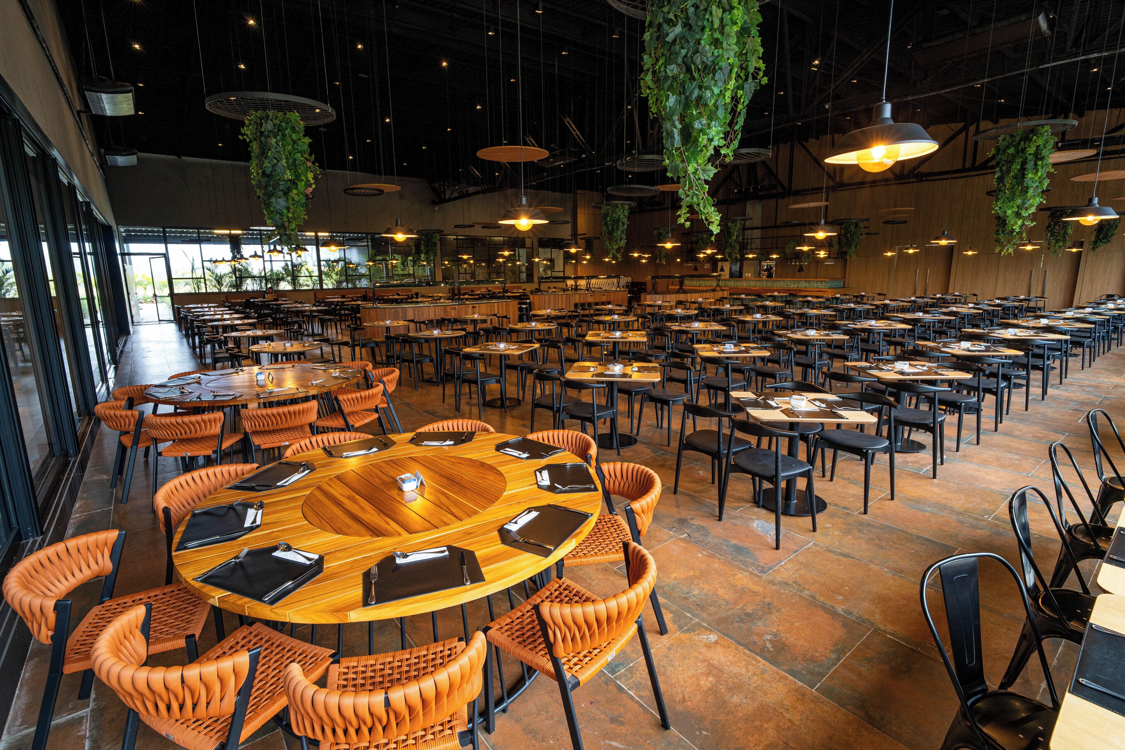 A spacious indoor dining area with multiple tables and chairs, large hanging plants, and a high ceiling.