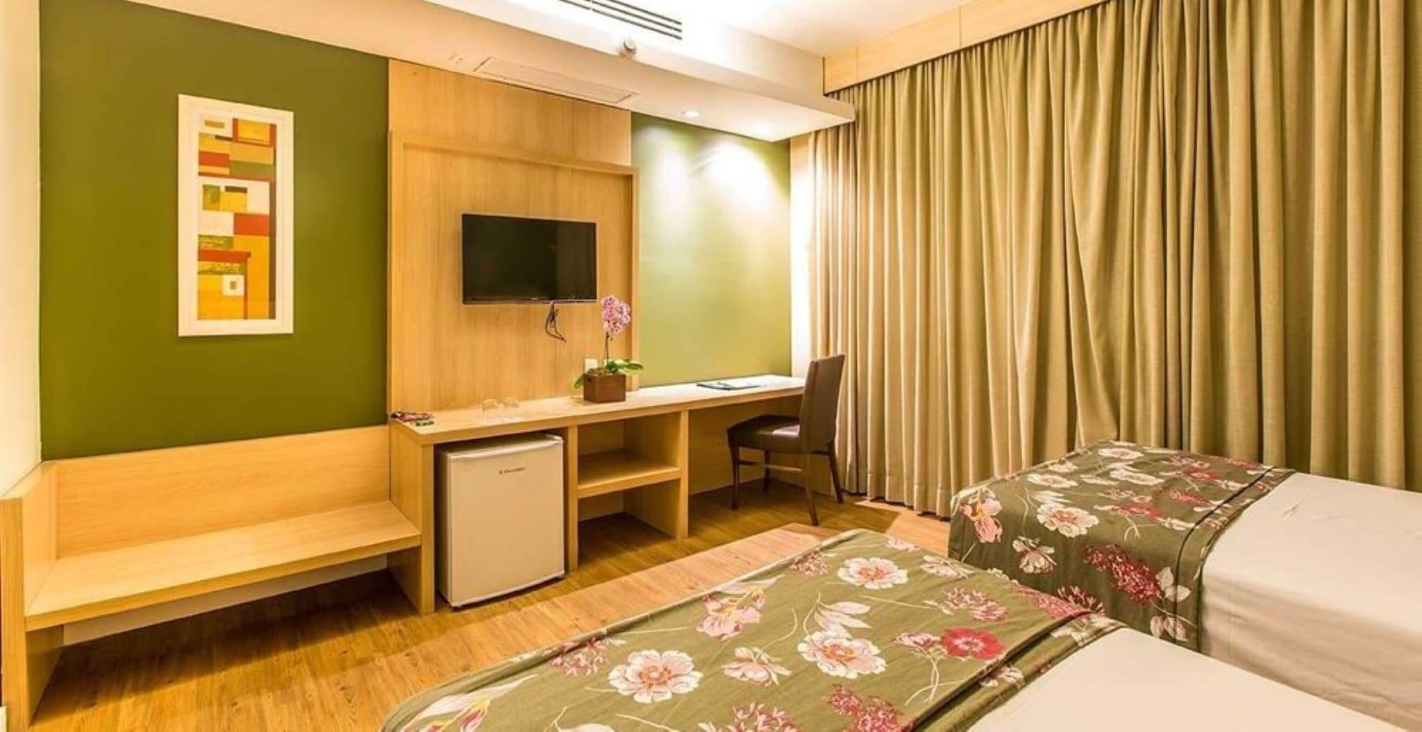 A hotel room with a desk, chair, two beds, a small refrigerator, and a TV.